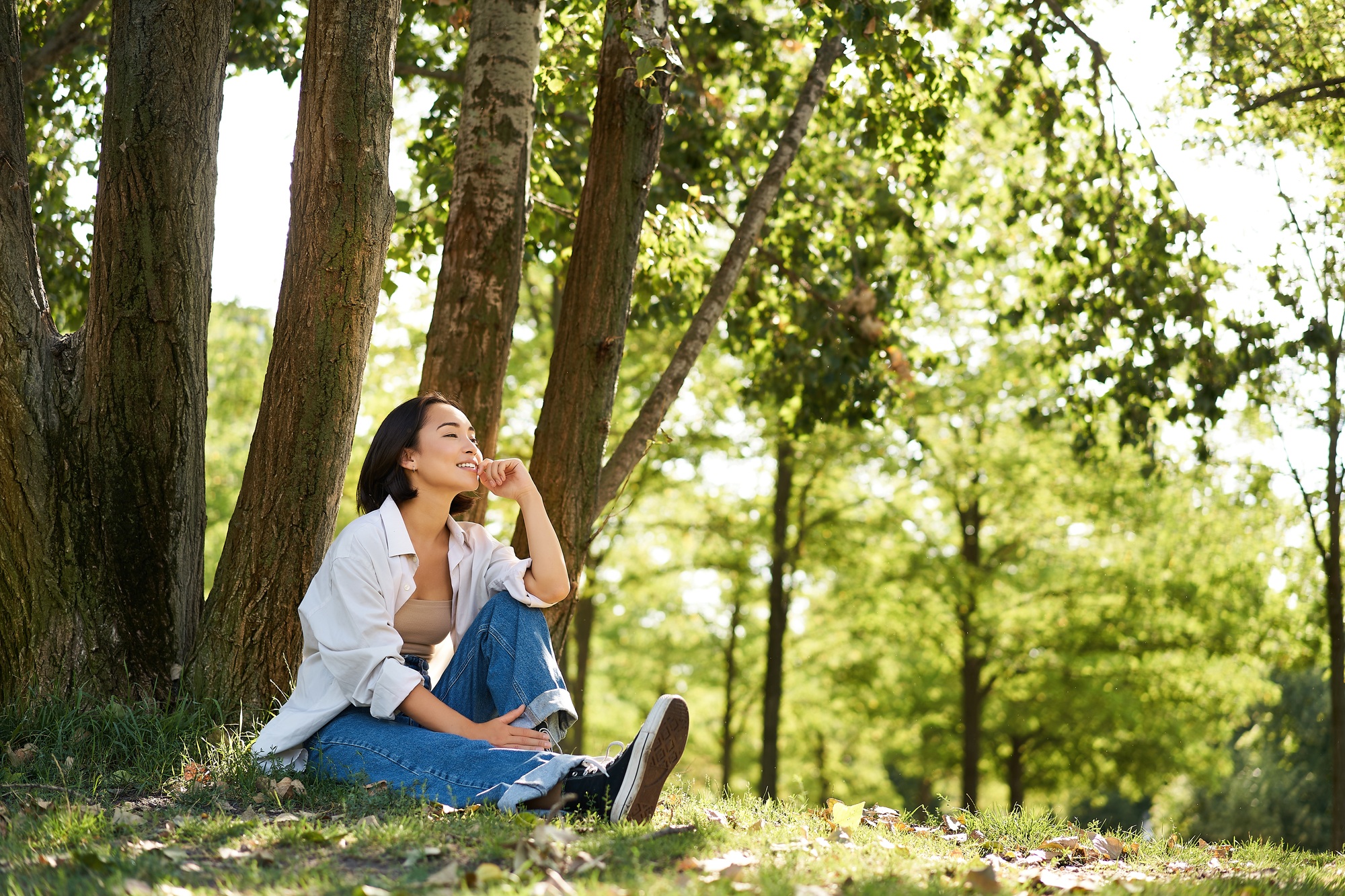 Relaxed young woman, resting near tree, sitting in park on lawn under shade, smiling and looking